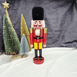 May include: A wooden nutcracker figure wearing a tall black hat, a red jacket with gold accents, yellow trousers, and black boots. The figure has a white beard, painted facial features, and stands on a round red base, accompanied by three small decorative trees.