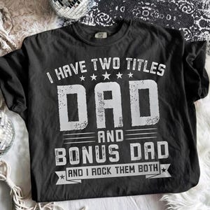 Può includere: T-shirt nero con testo bianco. Il testo recita "I HAVE TWO TITLES DAD AND BONUS DAD AND I ROCK THEM BOTH". Il testo è in grassetto, con un effetto invecchiato. La maglietta ha un girocollo e maniche corte.