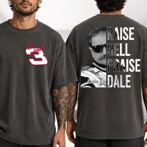 May include: Dark gray t-shirt featuring a white number 3 with a red outline and a cursive logo on the front. The back of the shirt displays a portrait of a man in sunglasses with the words "RAISE HELL PRAISE DALE" in white.