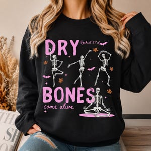 May include: A black long-sleeve sweatshirt featuring a graphic print with the text "DRY BONES came alive" and "Ezekiel 37:5". The design includes four white skeletons in various poses, pink bats, and orange autumn leaves on a black background.
