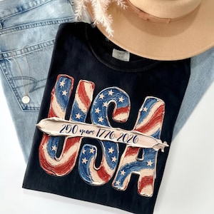 Può includere: T-shirt nero con un design patriottico USA in rosso, bianco e blu, con stelle e strisce. La maglietta include il testo "250 years 1776-2026". Un cappello color cachi e jeans blu sono anche nell'immagine.