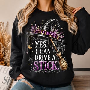 May include: A black long-sleeve sweatshirt featuring a graphic print of a witch hat, a broomstick, a spider web, a crescent moon, and a spider. The text reads, "YES, I CAN DRIVE A STICK." The design includes purple flowers and gold accents.