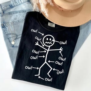 May include: Black t-shirt with a white graphic of a stick figure surrounded by the word "ow!" in a distressed font. The shirt is folded on top of a pair of light blue jeans, a tan hat, and a gold necklace.