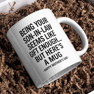 May include: White ceramic mug with black text that reads "BEING YOUR SON-IN-LAW SEEMS LIKE GIFT ENOUGH... BUT HERE'S A MUG - HAPPY MOTHER'S DAY". The mug is inside a gift box with brown paper filler.