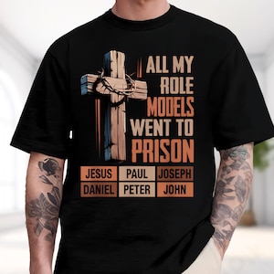 May include: A black short-sleeved t-shirt featuring a graphic of a wooden cross with a crown of thorns. The text reads: ALL MY ROLE MODELS WENT TO PRISON, followed by a grid of names: JESUS, PAUL, JOSEPH, DANIEL, PETER, and JOHN in orange boxes.