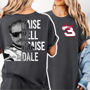 May include: Dark gray t-shirt featuring a black and white image of a man wearing sunglasses, with the text "RAISE HELL PRAISE DALE" in white. The right side of the shirt displays a red number 3 with a signature.