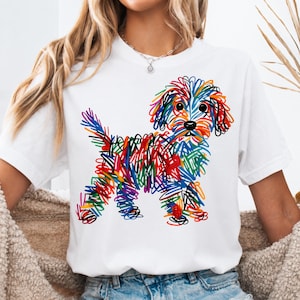 May include: A white short-sleeved t-shirt featuring a colorful graphic of a dog created with a scribbled, multi-colored line art style. The design incorporates vibrant shades of red, orange, yellow, green, and blue, forming the shape of a puppy.
