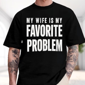 May include: A black short-sleeved t-shirt featuring white text printed on the chest that reads, MY WIFE IS MY FAVORITE PROBLEM. The shirt has a crew neckline and a standard fit, with the bold, capitalized lettering centered across the front.