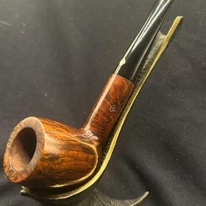 May include: A brown wooden tobacco pipe with a polished finish and a black stem, featuring a small diamond-shaped logo and text on the shank. The pipe rests in a curved, metallic gold-toned stand with a textured base and a smooth, angled support.