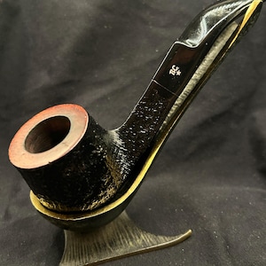 May include: A black tobacco pipe with a textured bowl and a smooth stem featuring the letters BC and a star symbol. The pipe rests in a metallic, dark-toned stand with a wide, curved base and a rounded cradle for the pipe bowl.