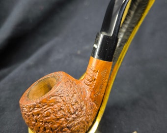 May include: A brown tobacco pipe with a textured bowl and a black stem, resting on a gold-colored stand. The pipe has a curved design and a smooth, polished finish. The stand is made of metal and has a unique shape.