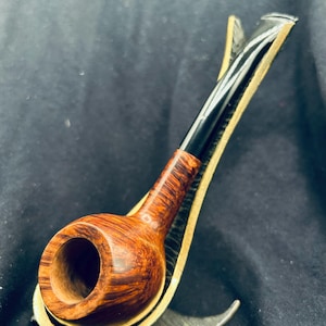 Könnte beinhalten: Eine Tabakpfeife aus Holz mit gebogenem Stiel und rundem Kopf. Die Pfeife hat eine dunkelbraune Oberfläche mit sichtbarer Holzmaserung. Sie ruht auf einem goldfarbenen Metallständer vor dunklem Hintergrund.