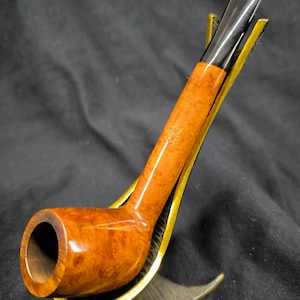 Pode incluir: Um cachimbo de madeira castanho com um bocal preto, apoiado num suporte de metal dourado. O cachimbo tem um padrão de grão de madeira natural.