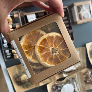 May include: A square cardboard box with a clear window, filled with dried orange slices. The box has a white label with the text "homestead on the hill". Other boxes and packages with dried fruit are in the background.