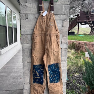 Puede incluir: Overol Carhartt marrón con correas marrones y parches azules en las rodillas. El overol está colgado de una percha verde. El logotipo de Carhartt es visible en el bolsillo delantero.