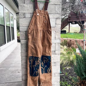 May include: Brown work overalls with adjustable straps and multiple pockets. The overalls have a patch of blue and white design on the knees. The overalls are hanging on a green hanger.
