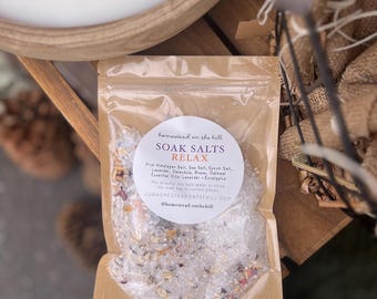 Puede incluir: Una bolsa transparente de sales de baño con la etiqueta "RELAX". La bolsa contiene una mezcla de sales, flores secas y avena. La etiqueta enumera ingredientes como sal del Himalaya, lavanda y eucalipto. La marca y el sitio web también son visibles.