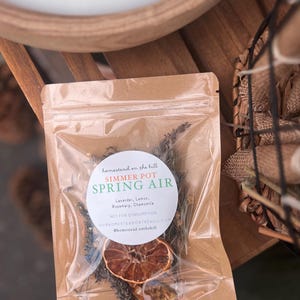May include: A clear plastic bag filled with dried herbs and a dried orange slice. The bag has a brown paper backing and a white label that reads "SIMMER POT SPRING AIR" with the ingredients listed below. The bag is on a wooden surface.