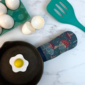 May include: A black cast iron skillet with a blue and white mushroom patterned oven mitt. A white plastic fried egg is in the skillet.