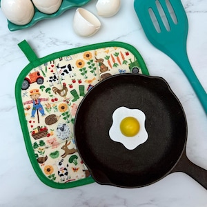 May include: A cast iron skillet with a fried egg, a farm-themed pot holder, and a teal spatula. The pot holder features a colorful pattern of farm animals, vegetables, and a scarecrow. White eggs and eggshells are in the background.