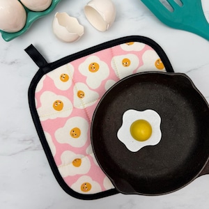 May include: A pink and white egg-themed pot holder with a black trim sits next to a cast iron skillet with a fried egg-shaped decoration. The pot holder has a repeating pattern of fried eggs. A teal spatula and a carton of eggs are in the background.
