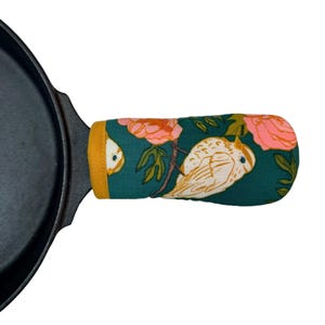 May include: A cast iron skillet with a decorative handle cover. The handle cover is teal with a floral and bird pattern in pink, yellow, and white. The edge of the handle cover is yellow.