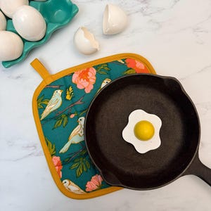 May include: A black cast iron skillet with a fried egg-shaped magnet. A teal floral patterned pot holder with birds and flowers is next to the skillet. White eggs are in a teal egg holder, and two eggshells are nearby.