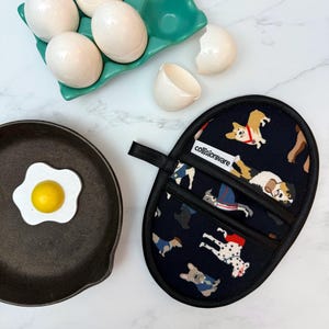May include: Black oven mitt with a dog pattern, next to a cast iron skillet with a fried egg, a teal egg carton with eggs, and an eggshell. The mitt has a black loop for hanging and the text "collisionware".