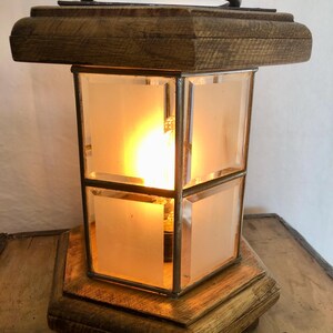 May include: A vintage lamp with a wooden base and top, featuring a rectangular glass enclosure with frosted panels. The lamp has a metal handle and a warm, glowing light source inside. The wooden components have a distressed finish.