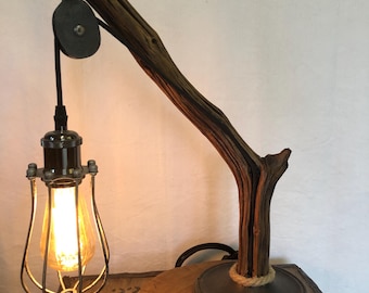 May include: A rustic desk lamp featuring a wooden branch as the main support. The lamp includes a metal pulley system and a caged lightbulb. The base is made of wood and metal, with a rope detail. The lamp has a warm, inviting glow.
