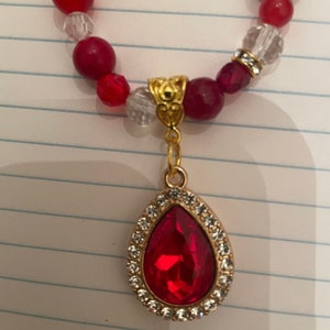 May include: A red gemstone pendant necklace with a gold chain and a teardrop-shaped red crystal surrounded by clear rhinestones.
