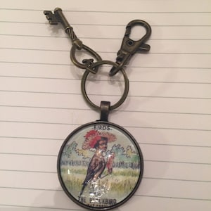 Puede incluir: Un llavero de bronce con un charm redondo de vidrio que presenta una ilustración vintage de un pájaro con el texto "BIRDS - THE BLACKBIRD - BY FRANK"