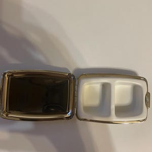 May include: A rectangular metal pill case with a hinged lid, featuring a polished silver-tone exterior. The interior contains a reflective mirror on one side and a white plastic insert with two separate compartments on the other side.