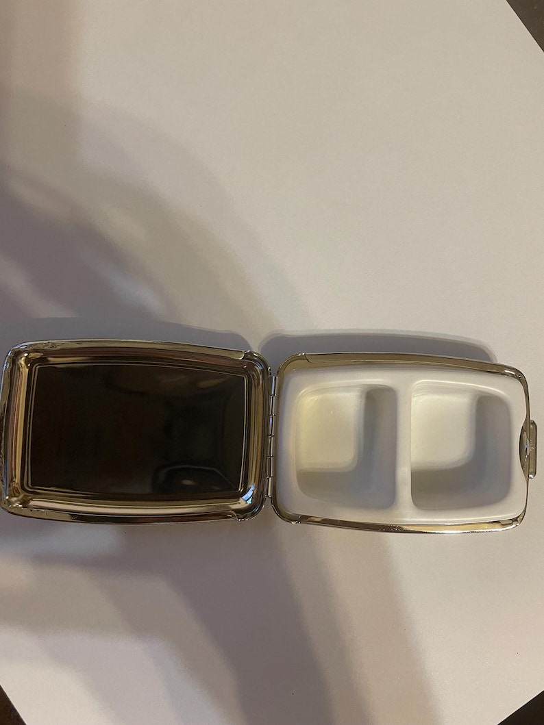 May include: A rectangular metal pill case with a hinged lid, featuring a polished silver-tone exterior. The interior contains a reflective mirror on one side and a white plastic insert with two separate compartments on the other side.