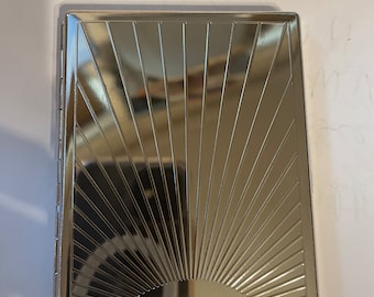 May include: A rectangular, silver-colored cigarette case. The case features a sunburst design on the front cover, with etched lines radiating outwards from a semi-circular base. The case is closed and has a hinged side.