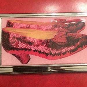 May include: A silver business card holder with a pink background and a pair of red sequined shoes with black accents.