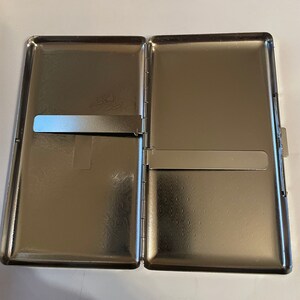 May include: A silver metal cigarette case with a hinged lid and a slide-out compartment. The case has a textured pattern on the outside.