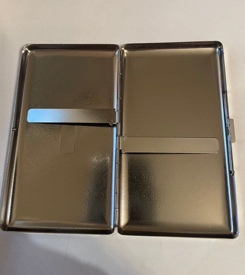 May include: A silver metal cigarette case with a hinged lid and a slide-out compartment. The case has a textured pattern on the outside.