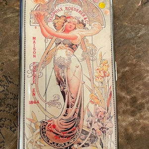 May include: A silver cigarette case with a vintage art nouveau design featuring a woman holding a tray of champagne glasses. The text on the case reads "Champagne Theophile Roederer & Co. Maison Fondée en 1864".