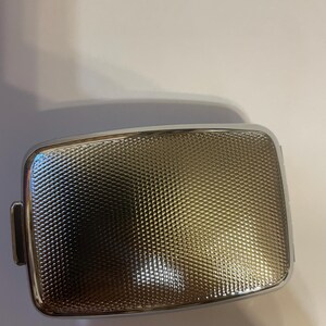 May include: A rectangular metal case with a polished silver-tone frame and a textured, cross-hatched surface pattern. The center panel features a fine, repeating geometric grid design that creates a subtle light-reflecting effect across the surface.