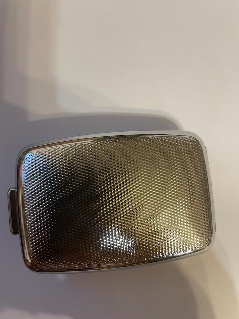 May include: A rectangular metal case with a polished silver-tone frame and a textured, cross-hatched surface pattern. The center panel features a fine, repeating geometric grid design that creates a subtle light-reflecting effect across the surface.