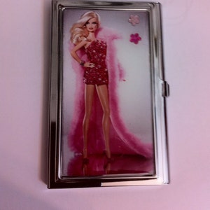 May include: A silver metal cigarette case with a pink and white image of a Barbie doll wearing a pink fur coat and a sparkly red dress. The doll is standing on a white background with pink flowers.