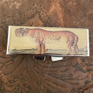 May include: A rectangular metal pill box with a hinged lid. The box features a vintage illustration of a leopard with brown spots on a cream-colored background. The leopard is standing on a grassy area.