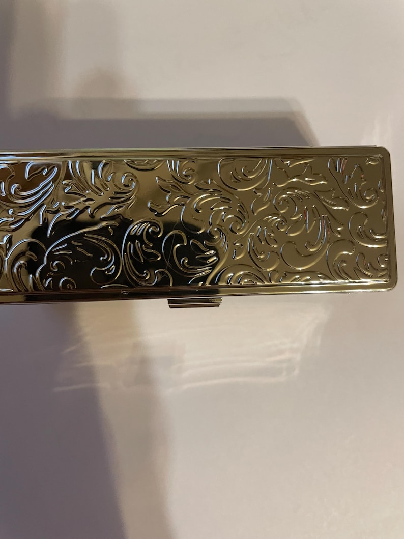 May include: A silver metal box with an ornate floral design. The box has a hinged lid and a clasp.