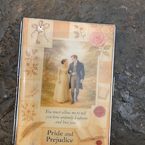 May include: A rectangular metal case featuring a scene from "Pride and Prejudice." The cover displays a couple walking in a garden, surrounded by floral and script elements. The text "You must allow me to tell you how ardently I admire and love you" is visible.