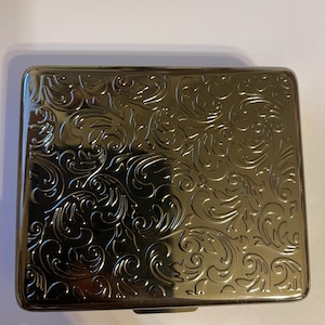 May include: A rectangular, gold-colored cigarette case with an ornate, swirling floral pattern. The case has a shiny, reflective surface and a secure closure. The design is a classic, vintage style.