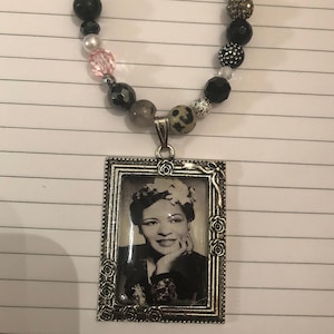 Op de afbeelding: Een zilverkleurige ketting met een hanger met een zwart-witfoto van een vrouw in een frame met een bloemenontwerp. De ketting is gemaakt met zwarte, witte en roze kralen.