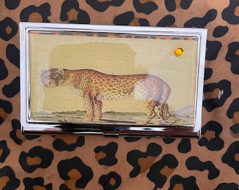 May include: A rectangular silver business card holder with a leopard illustration on a cream background. The leopard is brown with black spots. A small, gold-colored gem is set in the upper right corner. The background is a brown leopard print.
