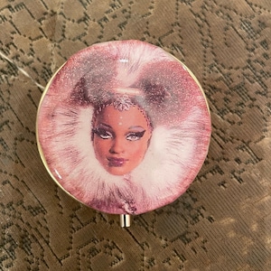 May include: A pink and white round compact with a picture of a woman with dark hair and dark eyes wearing a white fur collar. The woman is wearing a gold crown.