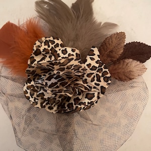 May include: A decorative hair accessory featuring a leopard print fabric flower, accented with brown and gray feathers and leaves. The accessory is set on a gray mesh base, creating a layered effect. This item is suitable for special occasions.